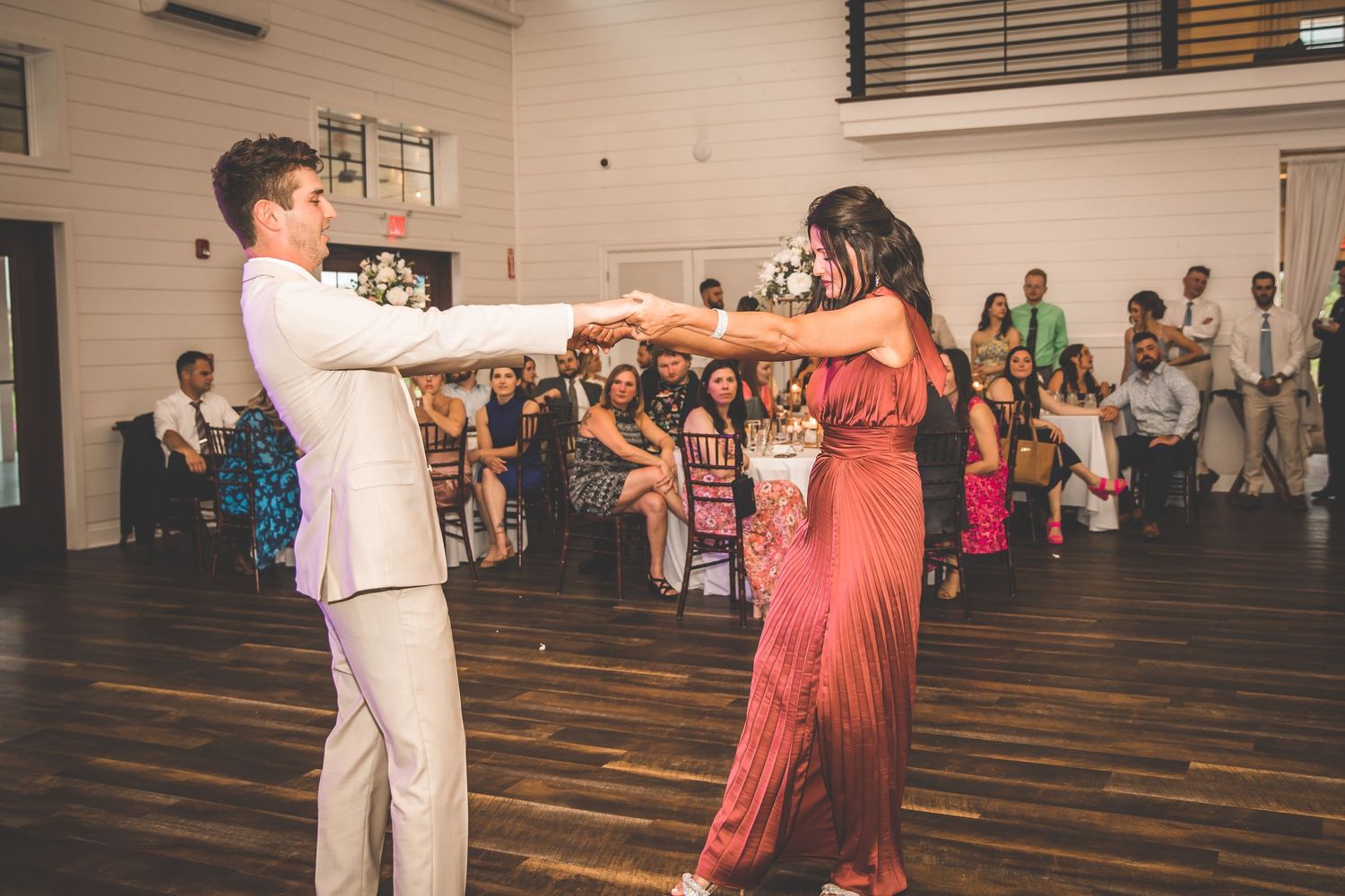 A dancing couple holds hands on a wooden dance floor while guests watch from the background.