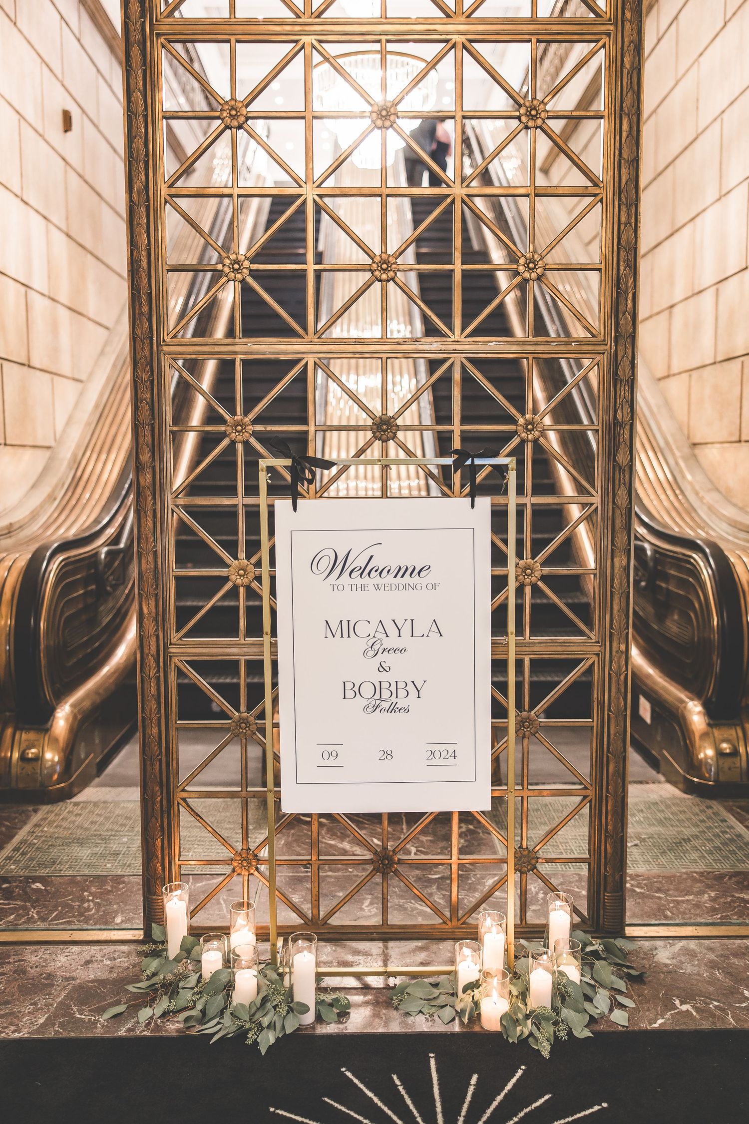 Elegant wedding welcome display with gold geometric frames and candles against a stone wall.