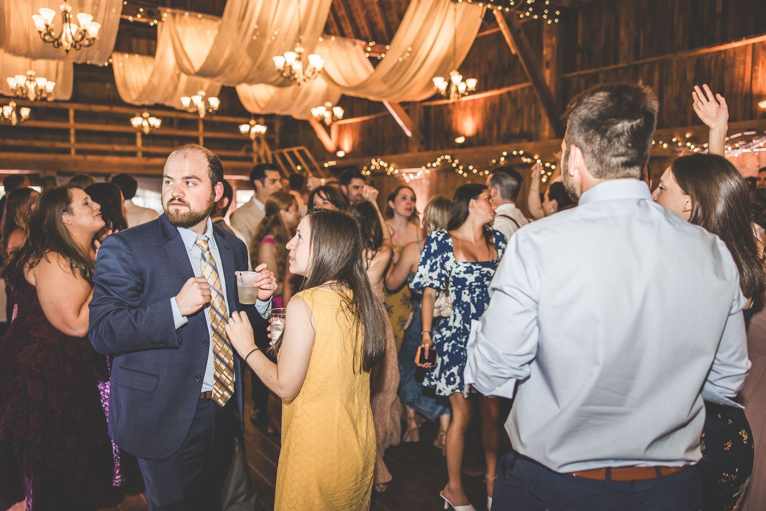 Wedding guests dancing and celebrating at lively barn reception with string lights.