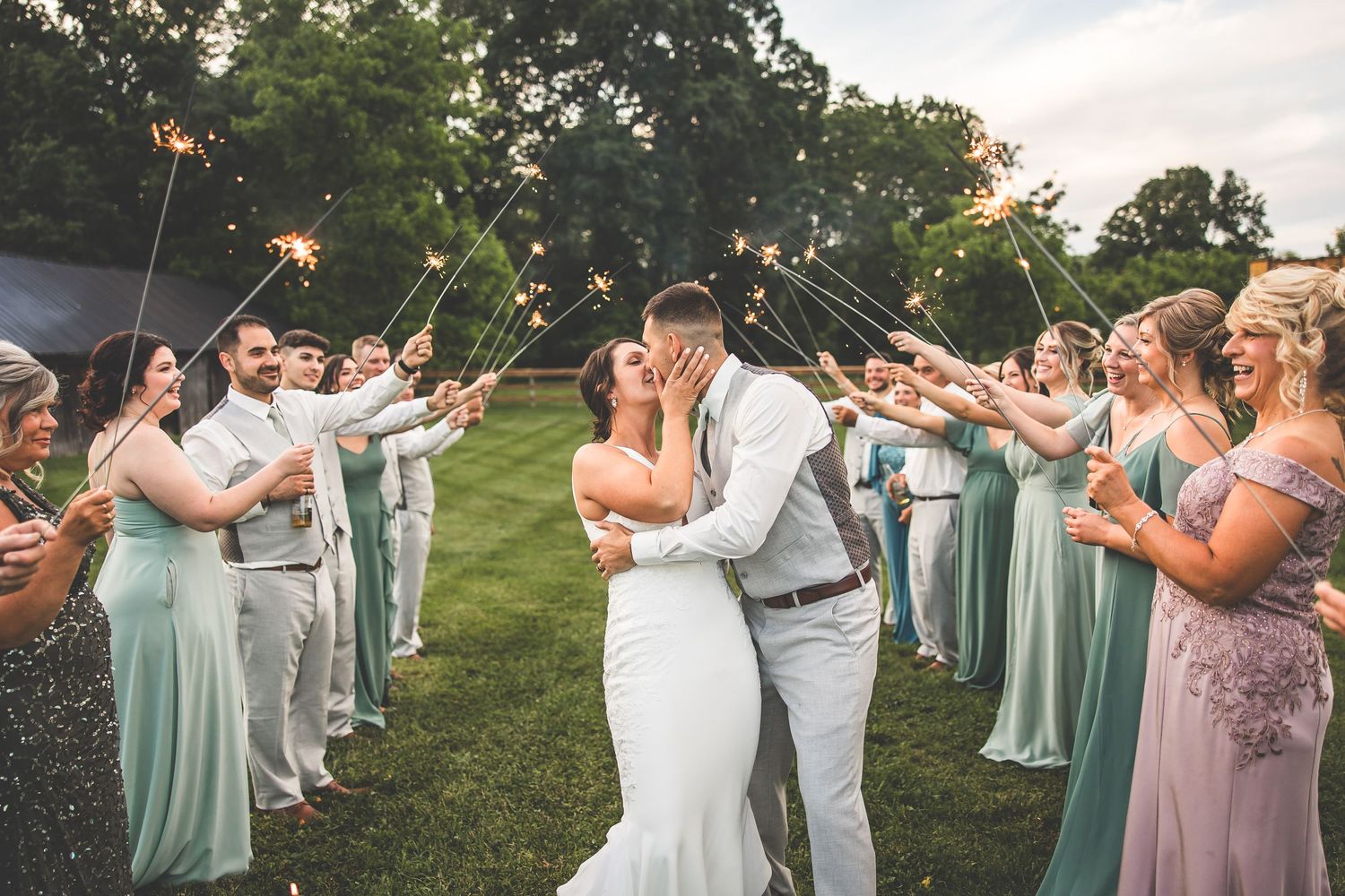 Bridal party in sage green dresses and gray suits dancing in a garden setting.