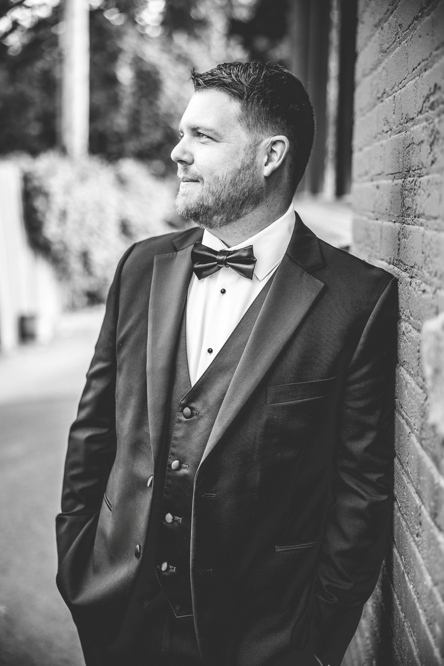 Black and white portrait of groom in tuxedo leaning against textured wall.