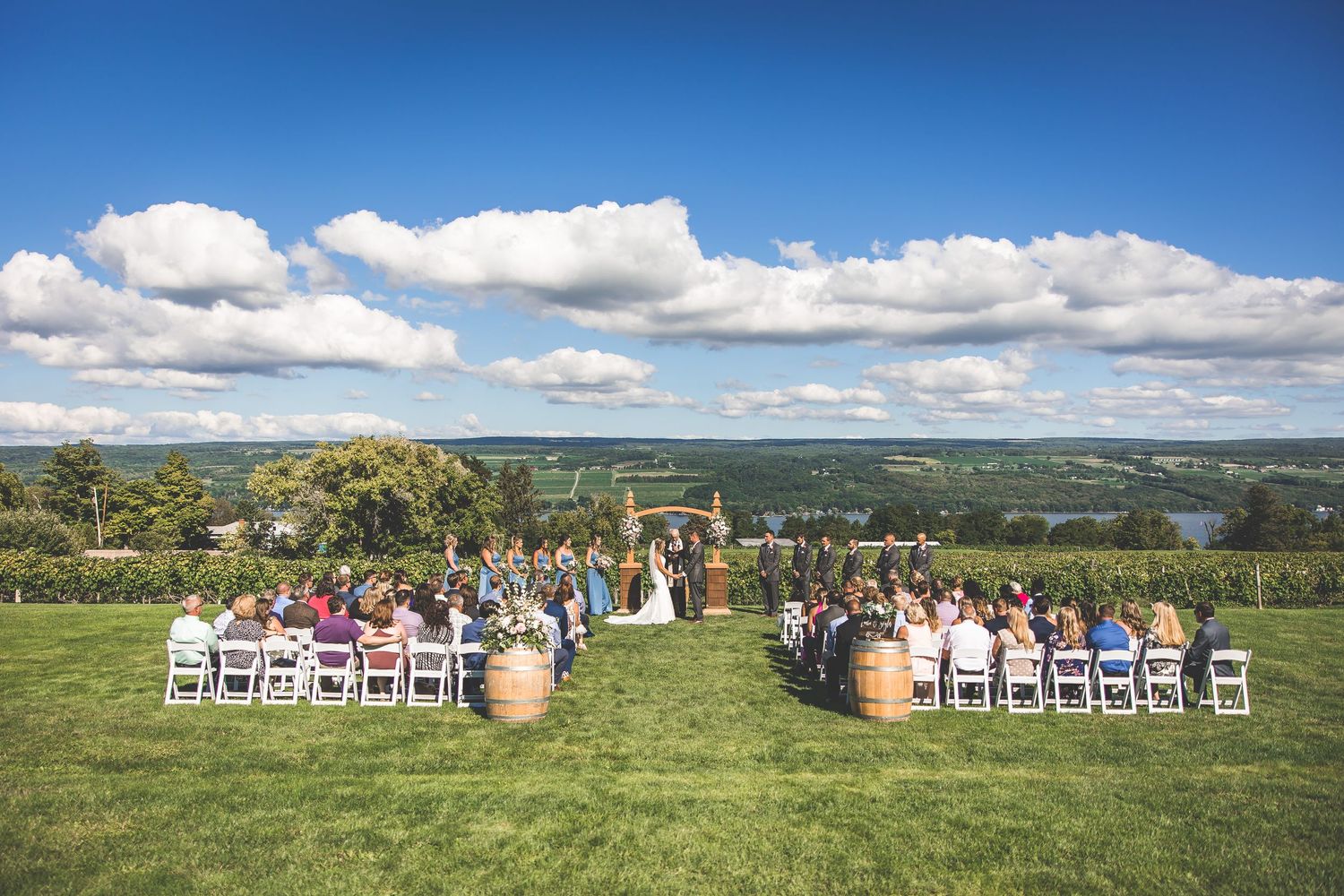 Panoramic view of a hilltop wedding ceremony with white chairs and wine barrels under blue skies.