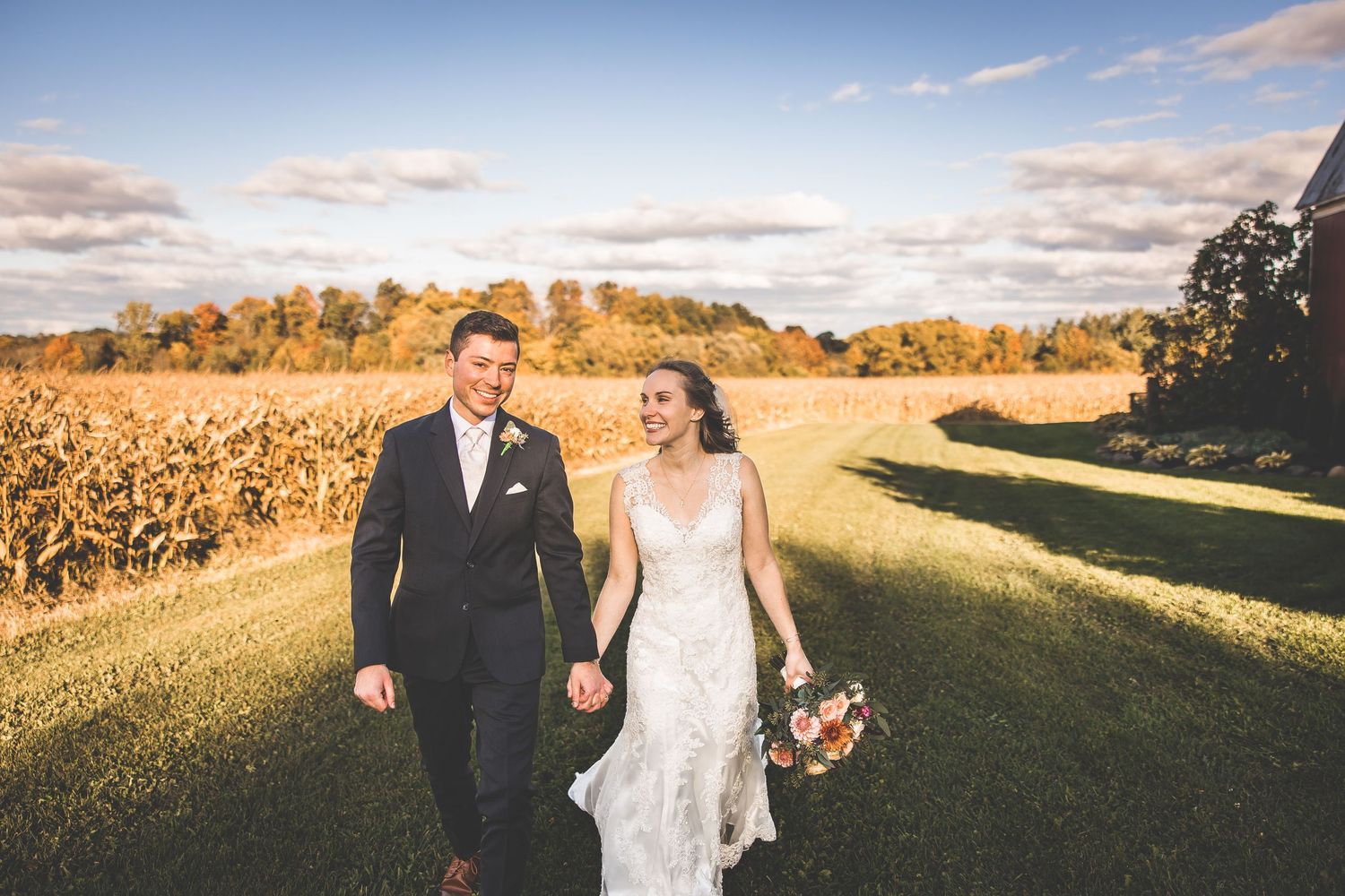 Couple walks hand in hand through golden field at sunset in wedding attire.