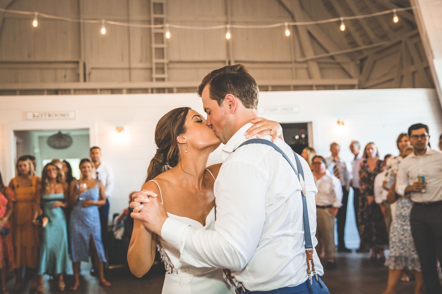 Wedding guests dancing and celebrating in a rustic indoor venue with string lights overhead.
