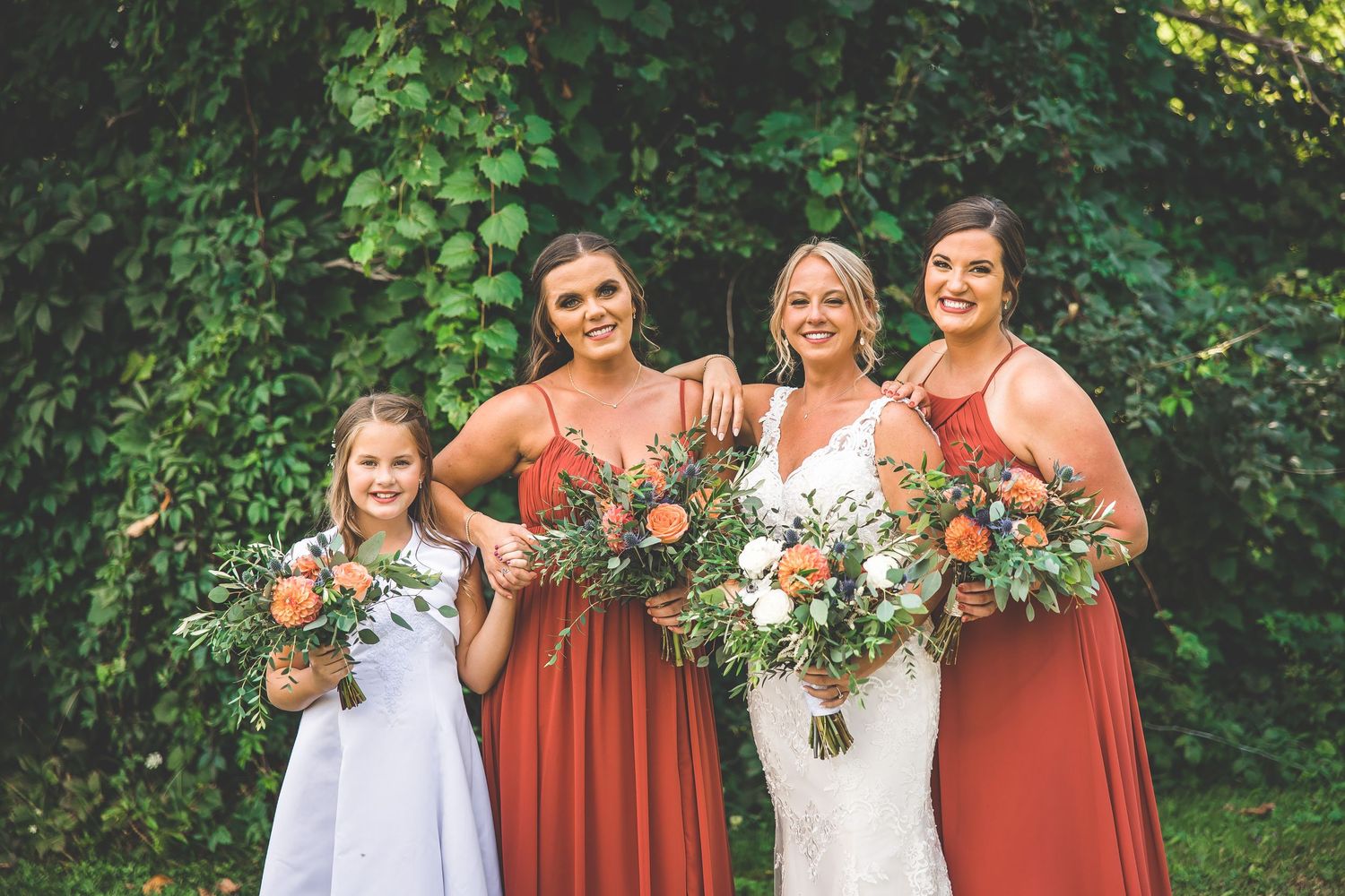 Bridal party wearing rust-colored dresses pose together outdoors with greenery backdrop.