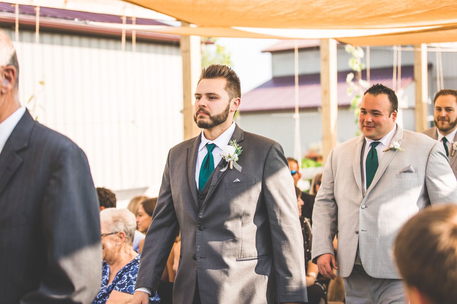 Groom walks down aisle in gray suit with teal tie under warm lighting.