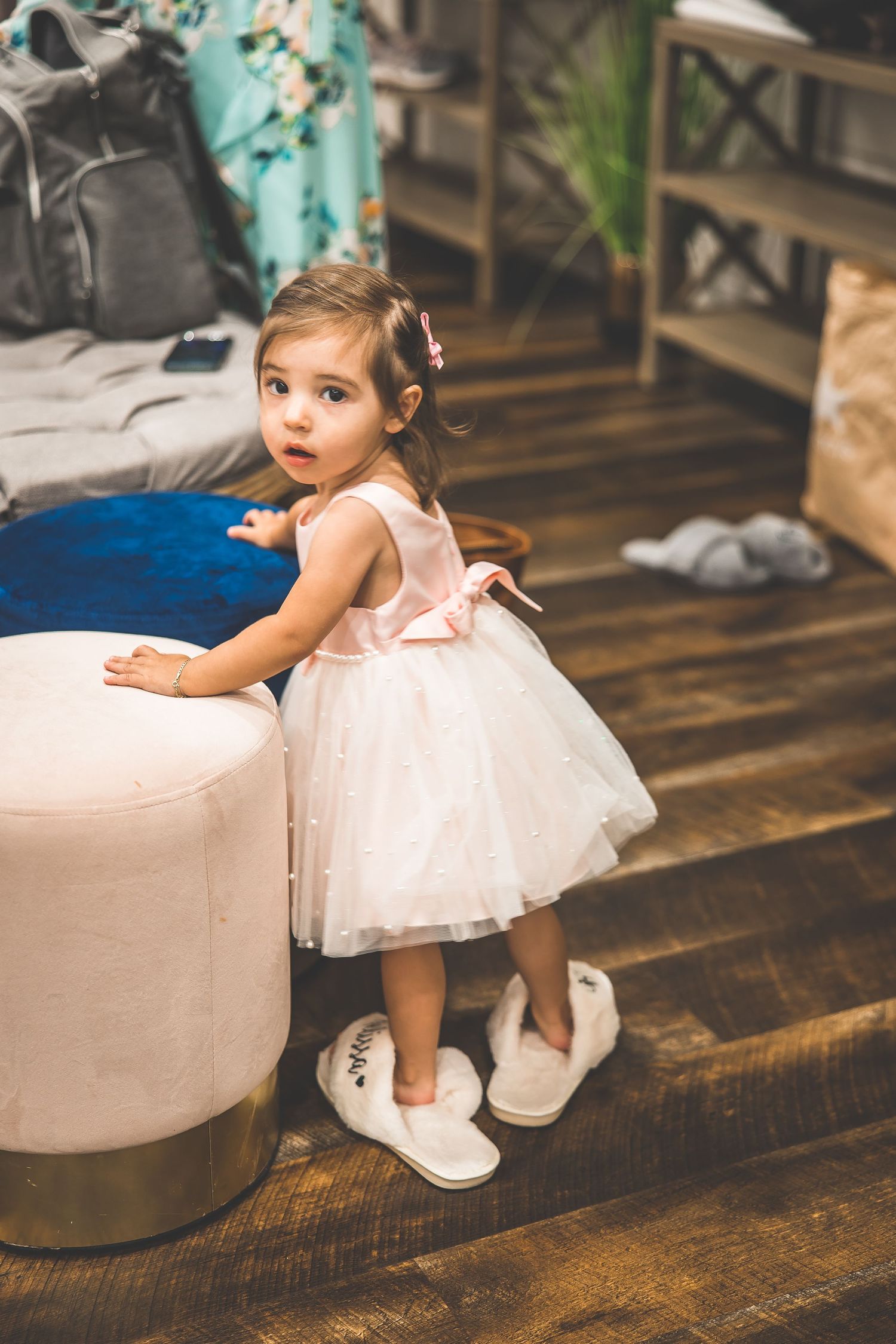 A young flower girl in a white tulle dress sits on a beige couch on hardwood floors.