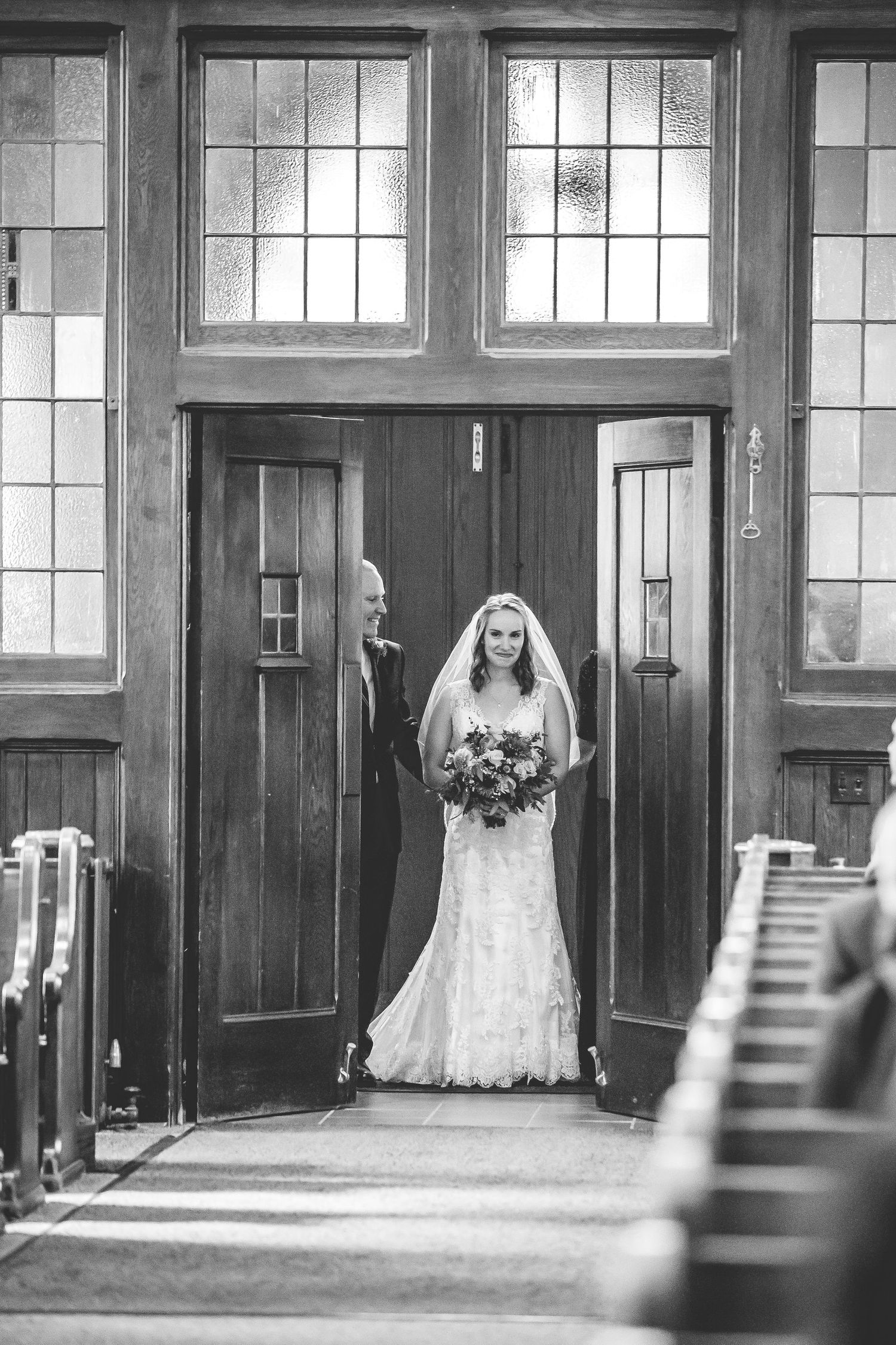 Black and white photo of a bride in a lace wedding dress standing in front of church windows.