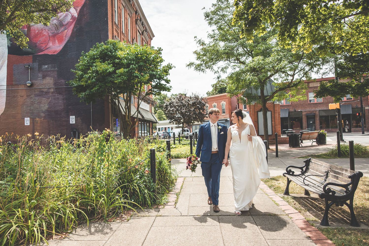 Couple strolling through a garden path with brick buildings and greenery in the background.