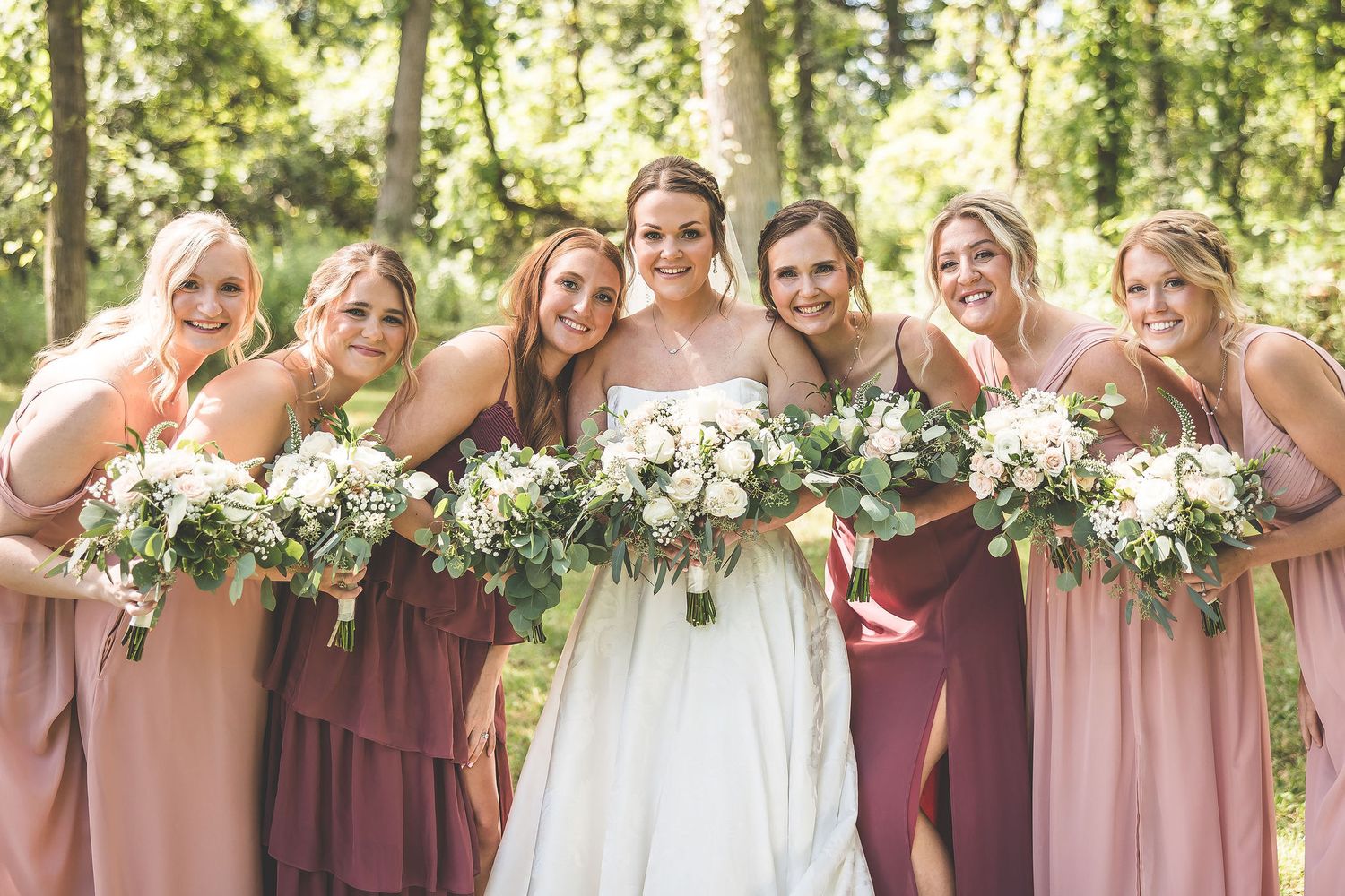 Bridesmaids in burgundy and blush pink dresses pose together with white floral bouquets in an outdoor setting.