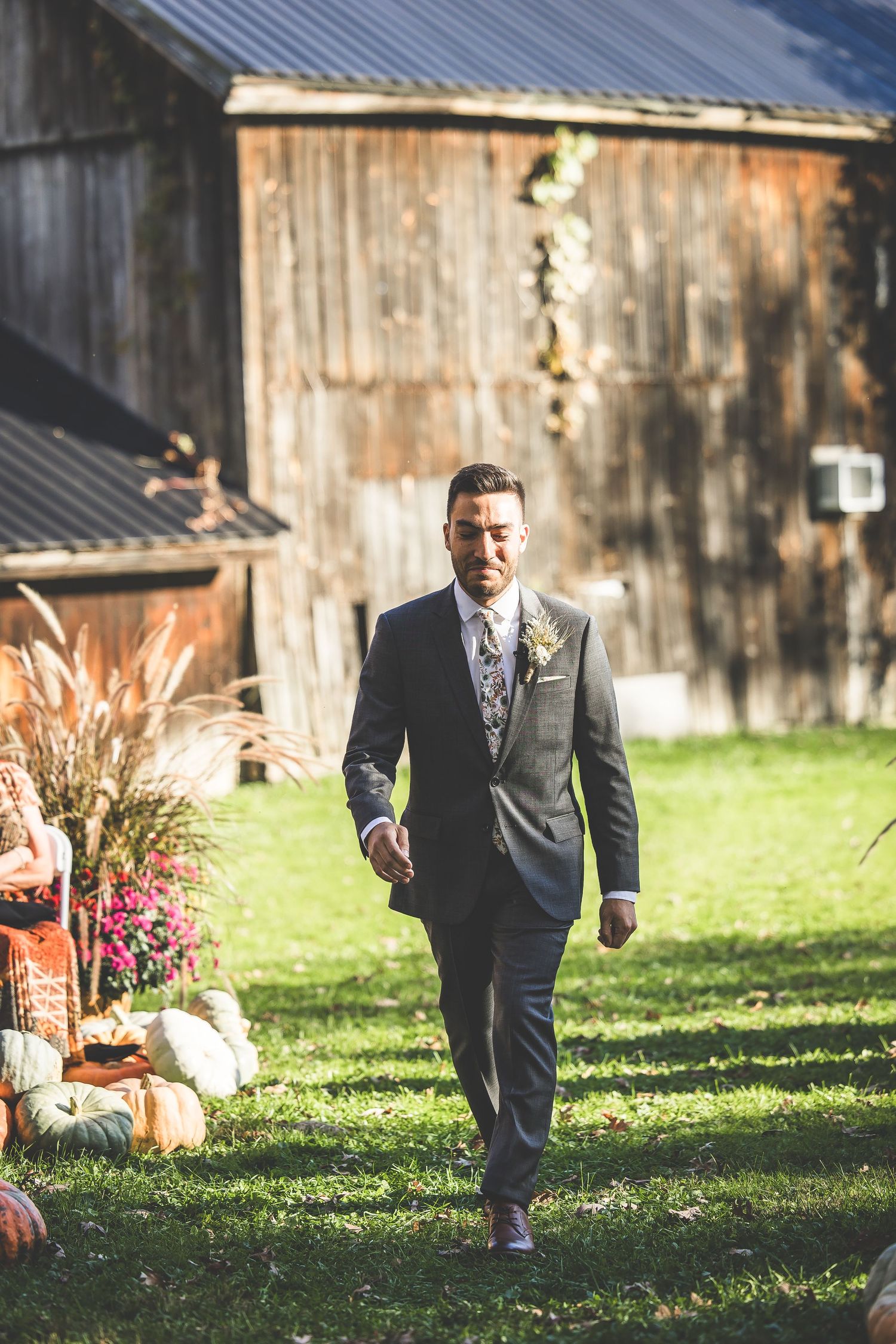 A groom in a dark suit walks along a rustic barn setting with fall decorations and pumpkins.