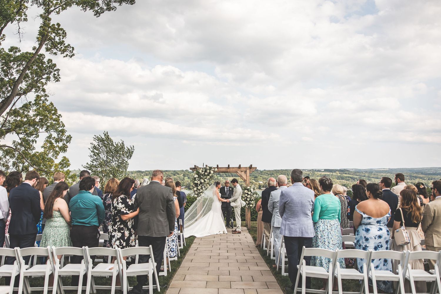 Rows of white chairs set up for an outdoor wedding ceremony with guests standing.