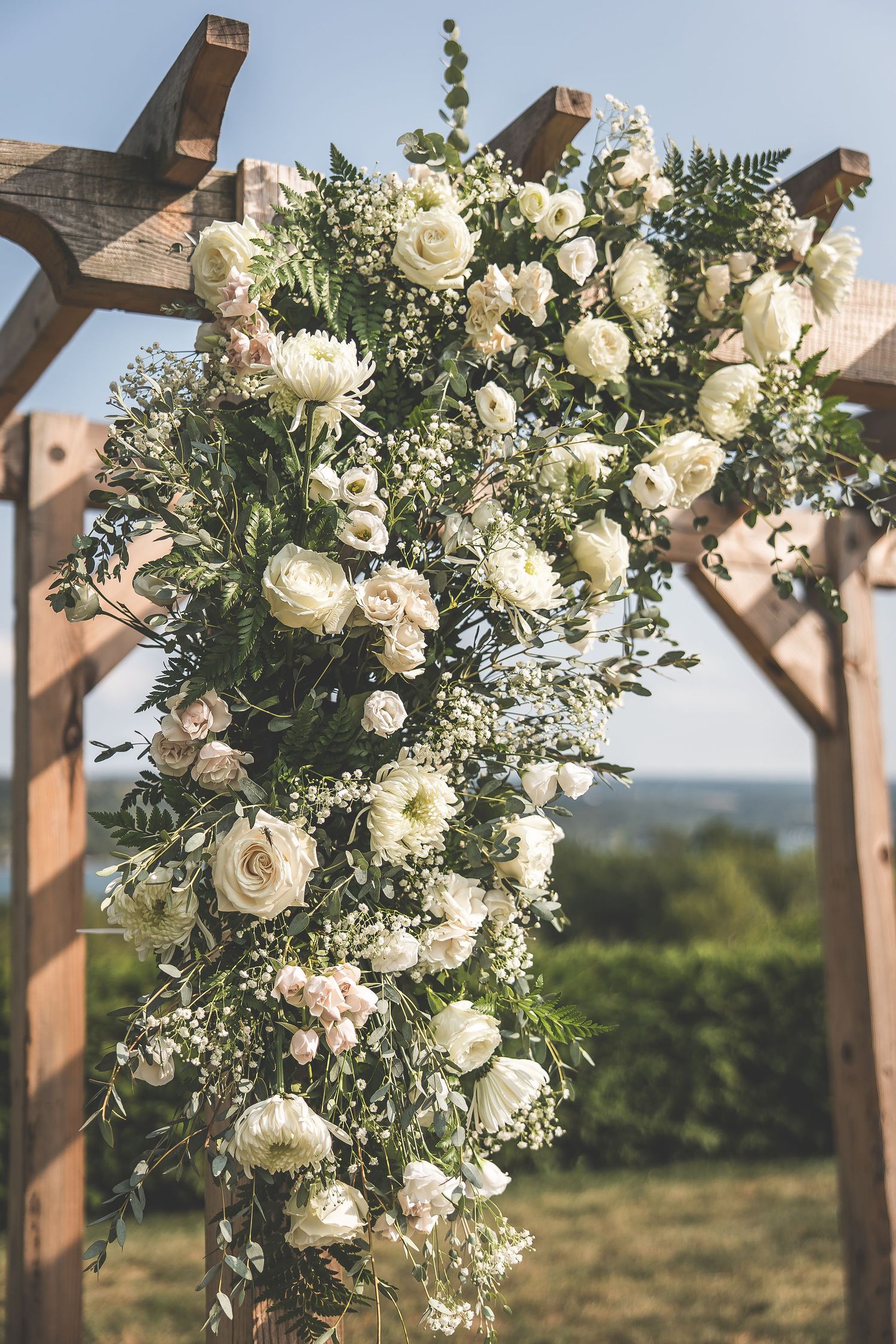 Wooden wedding arch decorated with cascading white roses and greenery against a scenic backdrop.