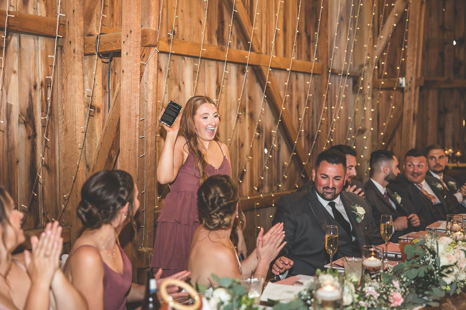 Wedding guests applauding during reception in rustic barn with string light decorations.