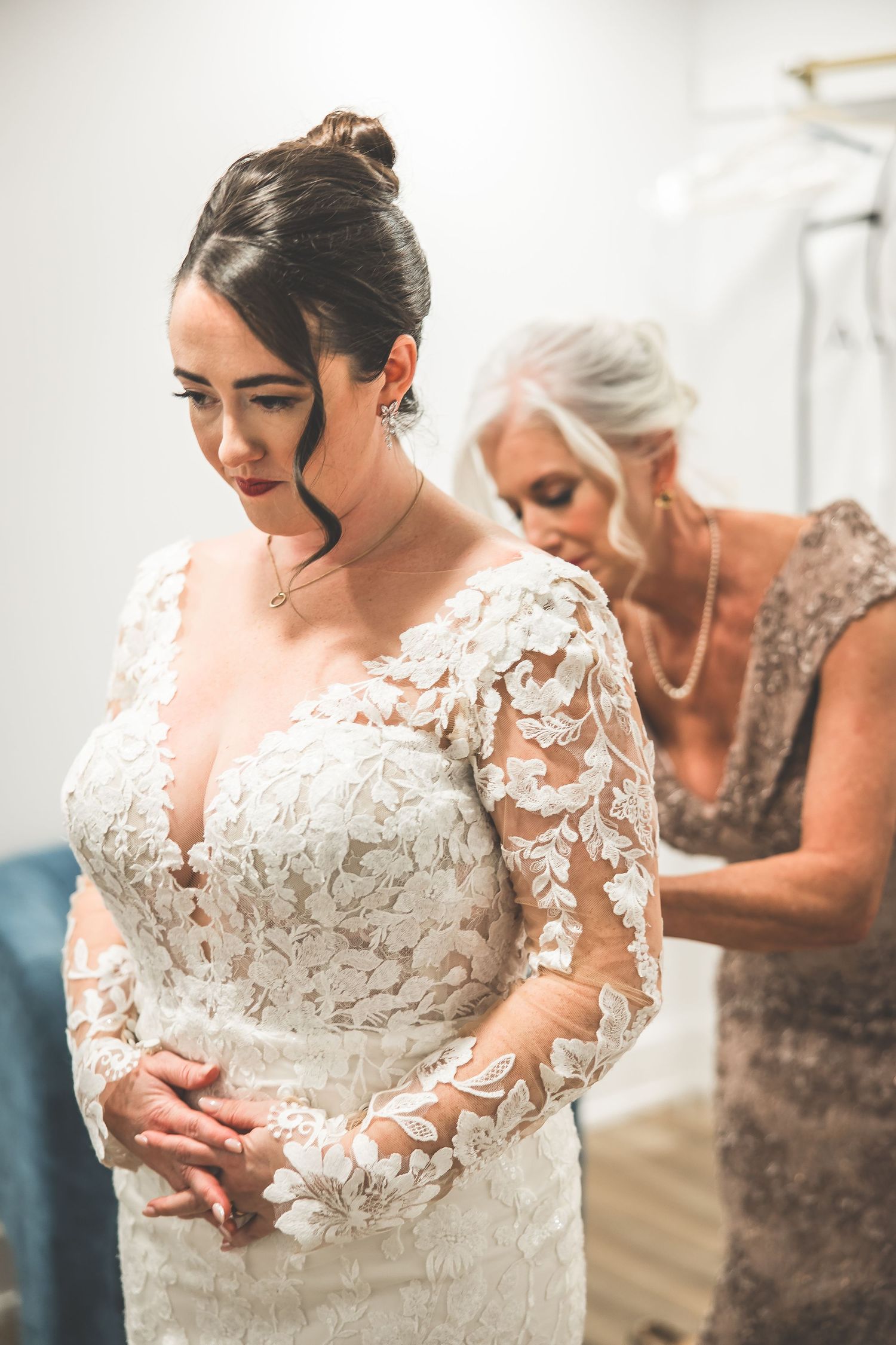 A stunning white lace wedding dress with intricate floral details and sheer long sleeves being fitted in a bridal suite.