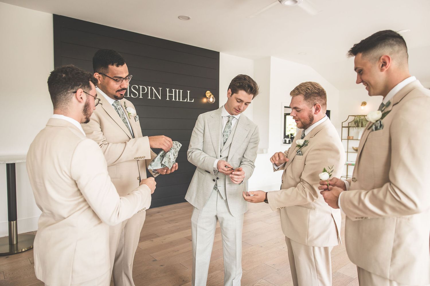 Groomsmen in matching beige suits preparing together in a bright modern indoor space.