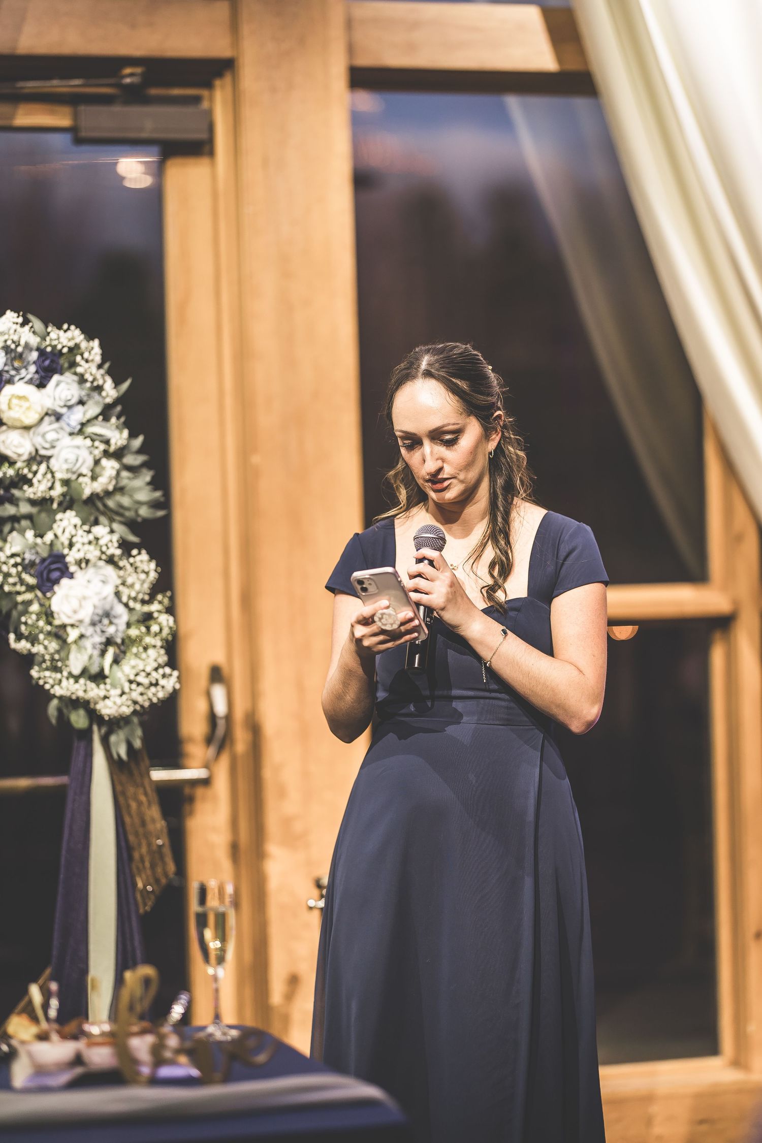 Speaker in navy dress giving toast during evening wedding reception near floral arrangements.