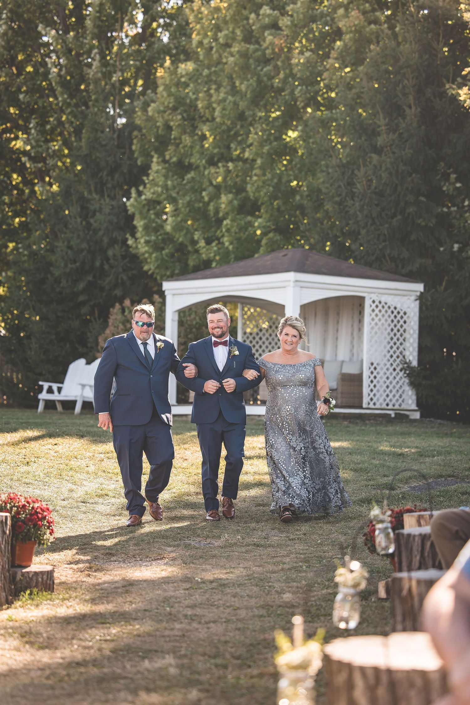 Wedding party members walk together on grassy hill with white gazebo in background.