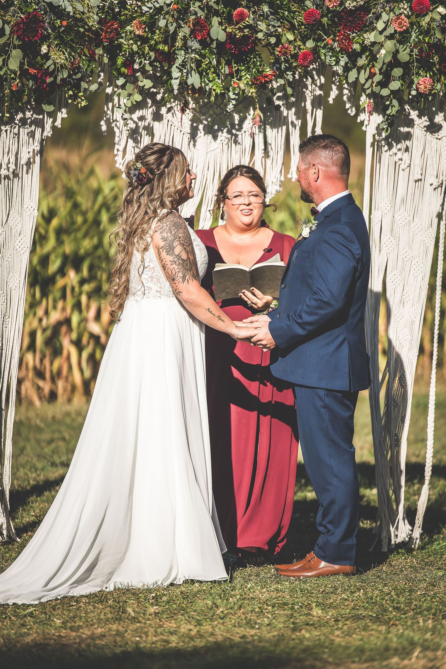Wedding ceremony with burgundy and navy color scheme under decorative macrame and floral arch.