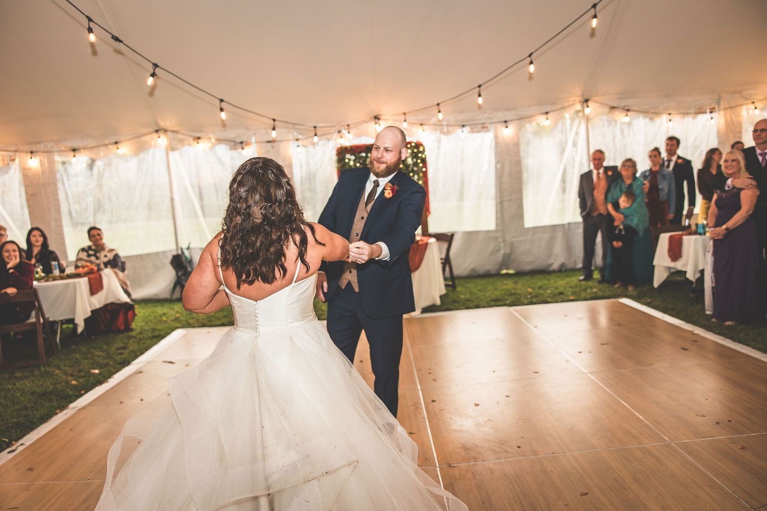 Elegant tent wedding reception with string lights and wooden dance floor surrounded by guests.
