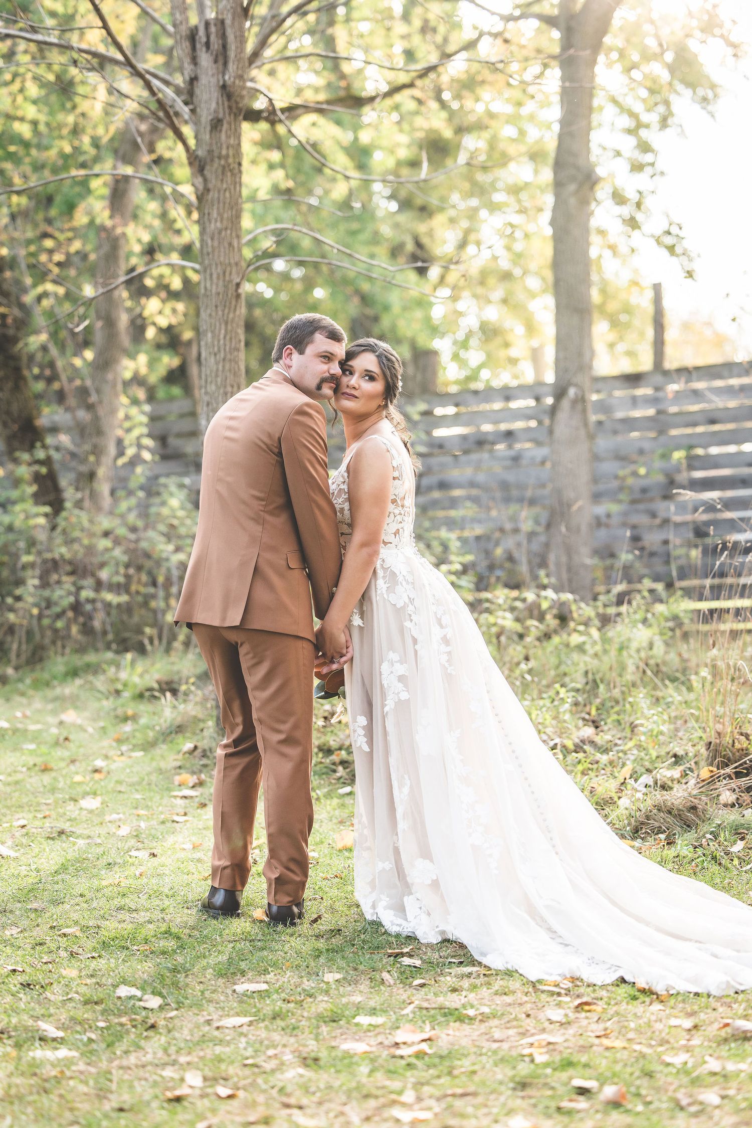 A romantic wedding couple embraces in a wooded outdoor setting with autumn foliage and sunlight filtering through trees.