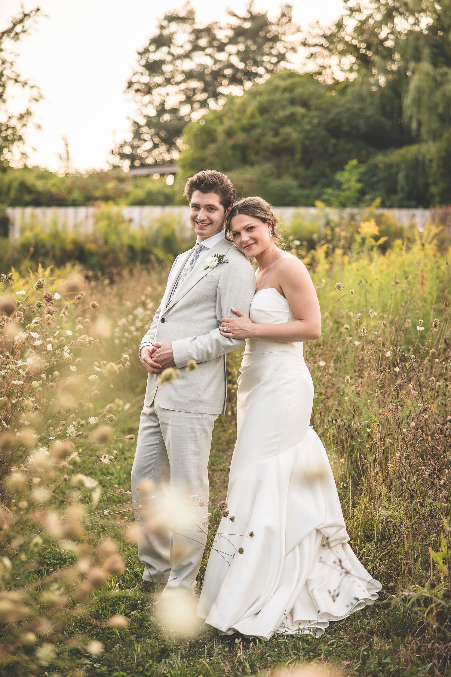 Newlyweds walk hand in hand through tall wildflowers with rustic barn in background.