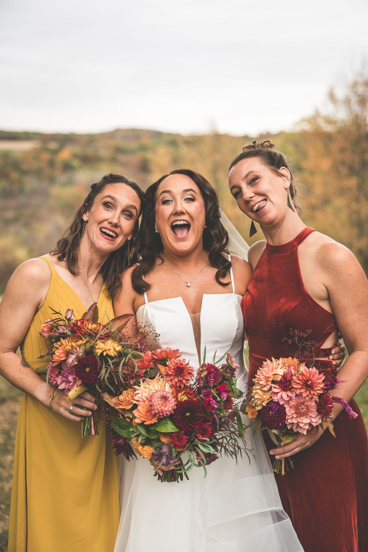 Wedding party members holding vibrant autumn bouquets laugh together in warm fall colors.