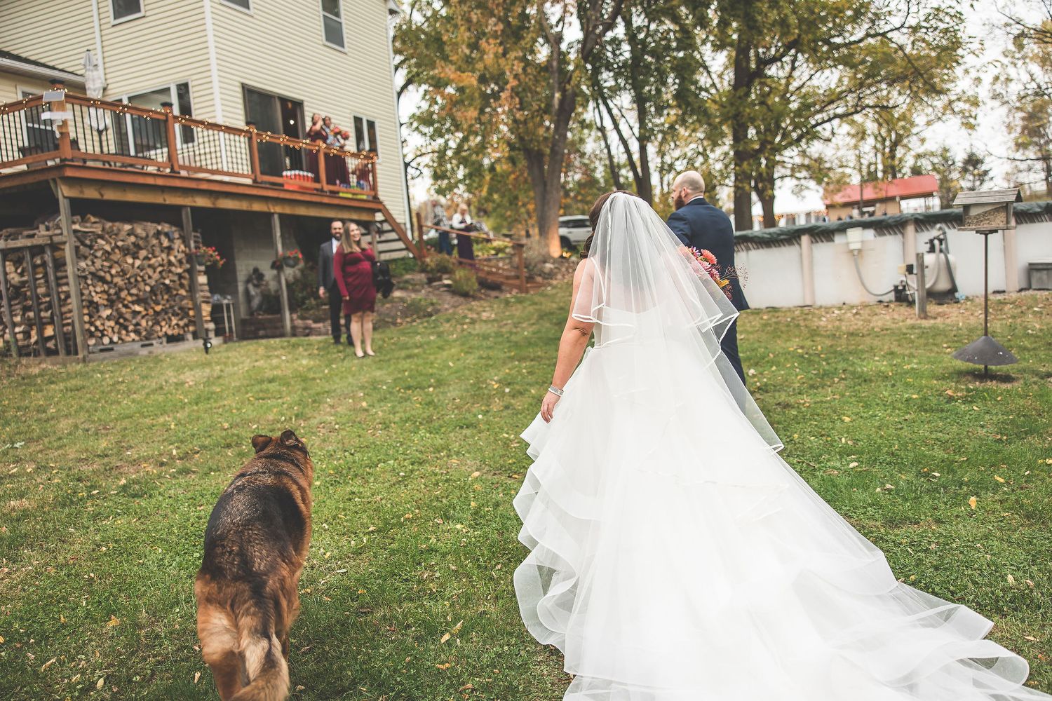 Wedding couple and their dog pose in front of a rustic house with fall foliage.
