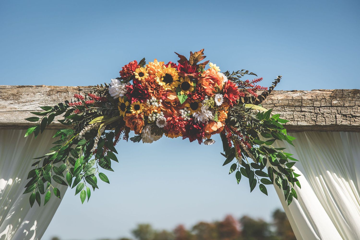 Wedding arch decorated with autumn flowers and white fabric against blue sky.