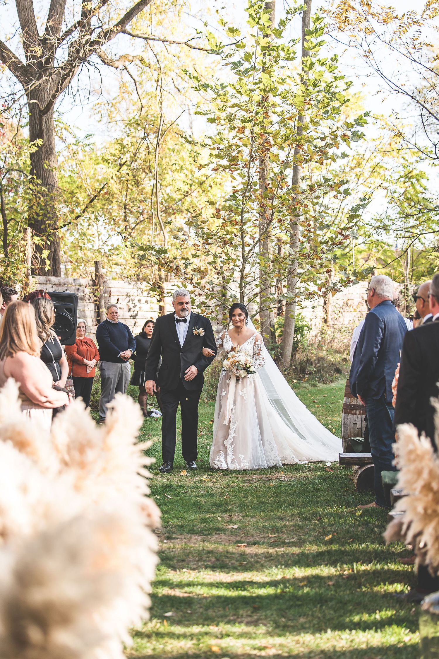 Outdoor wedding ceremony in a wooded setting with pampas grass decorations and gathered guests.