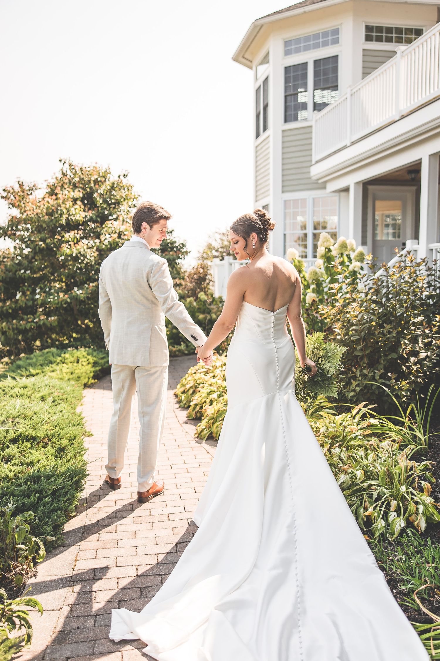 Bride and groom walking along garden path beside elegant coastal home at sunset.