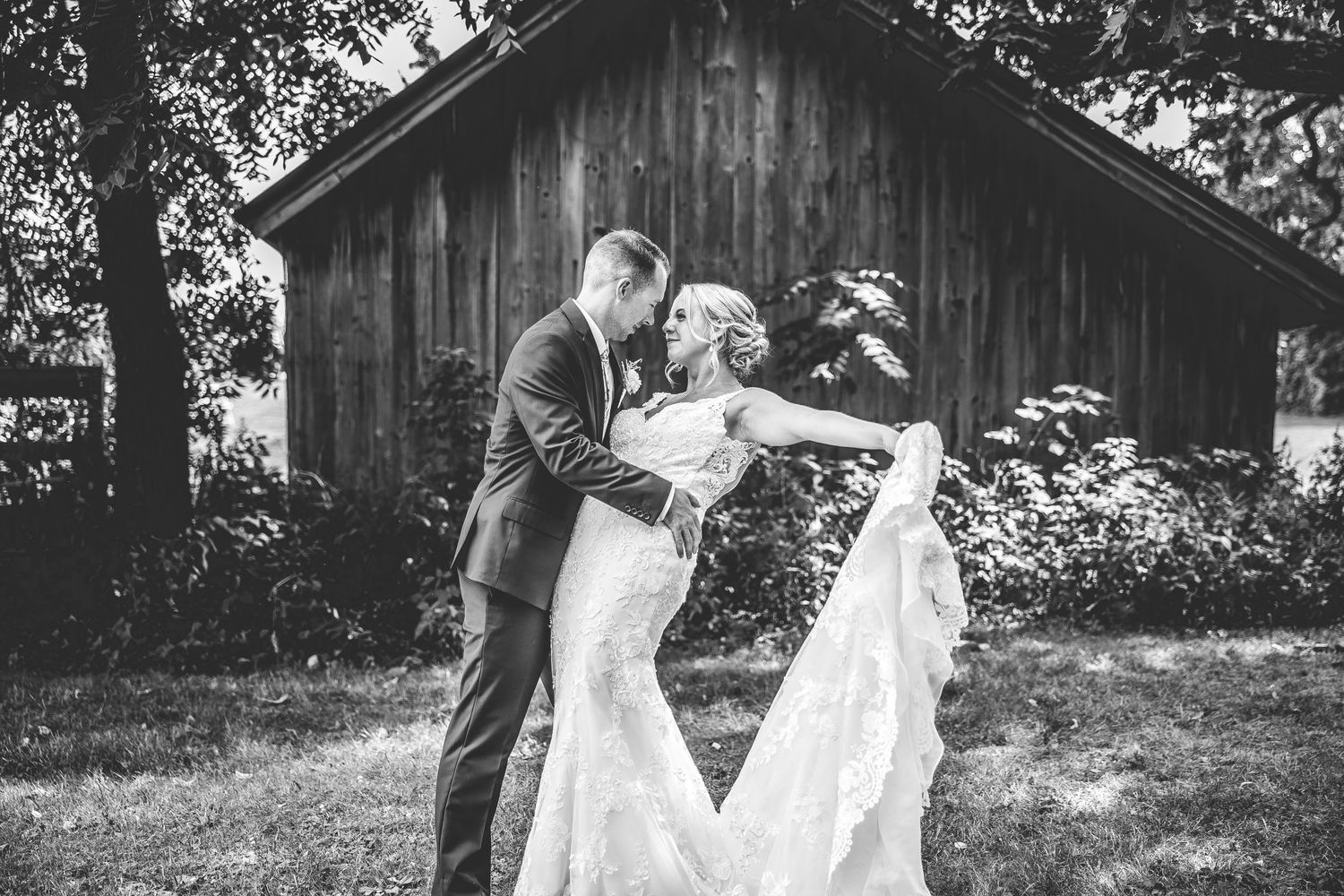 A black and white candid moment of newlyweds laughing together by a rustic barn.