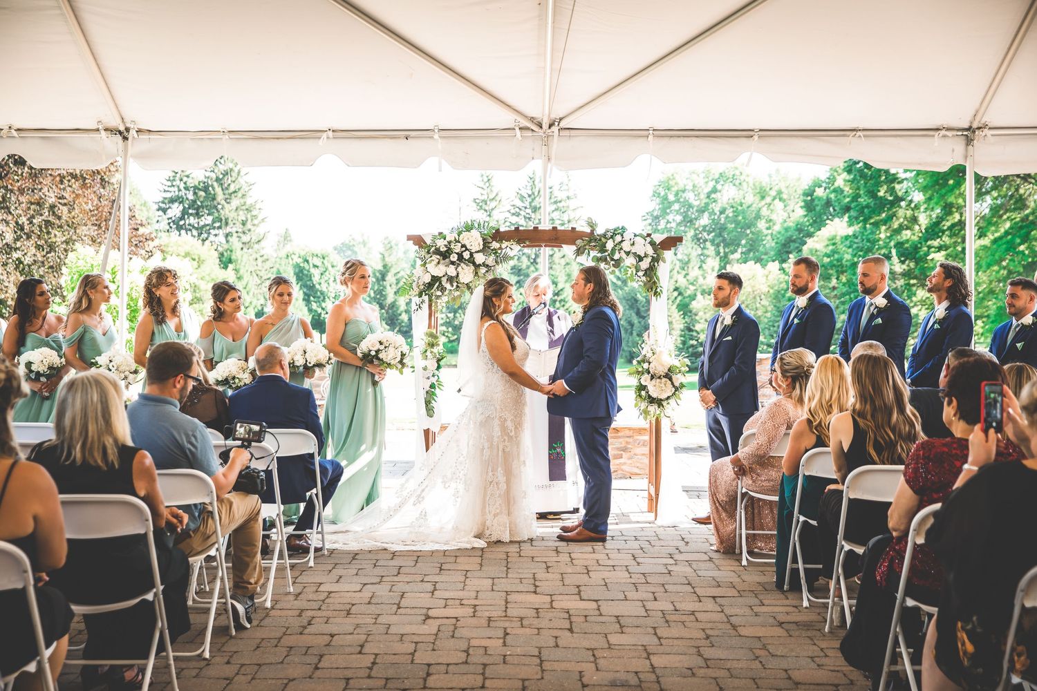 Side view of outdoor wedding ceremony under tent with guests seated on brick patio.