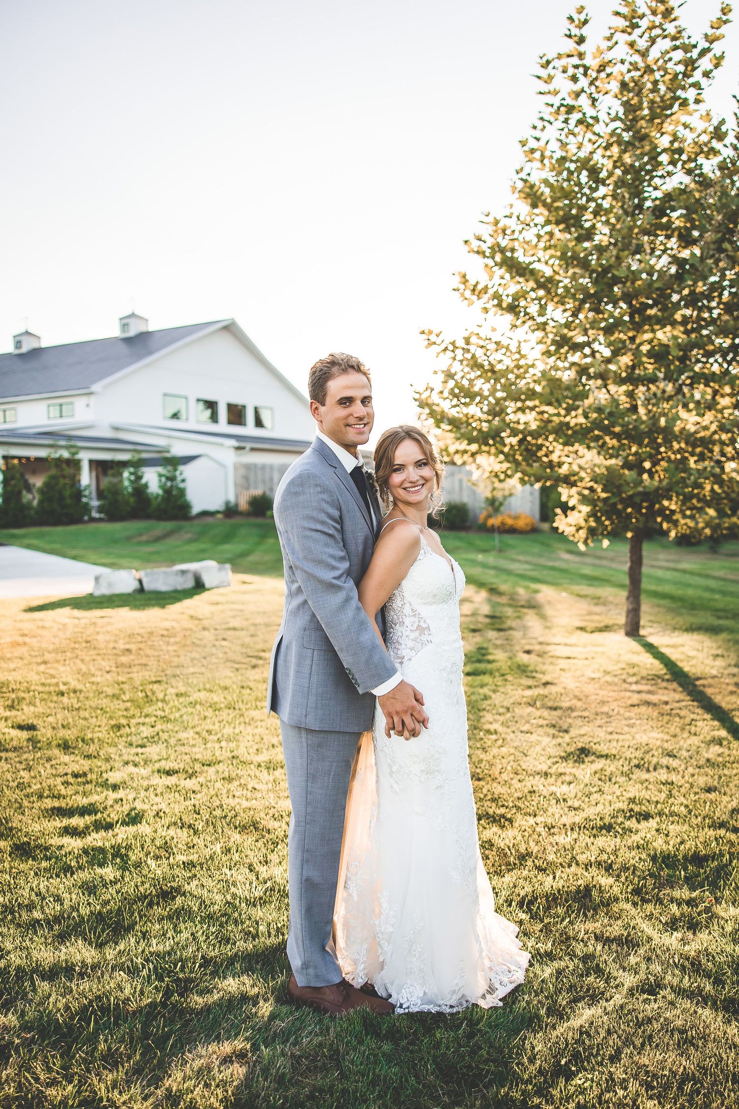 Newlyweds pose together on a sunlit lawn with a white farmhouse and tree in the background.
