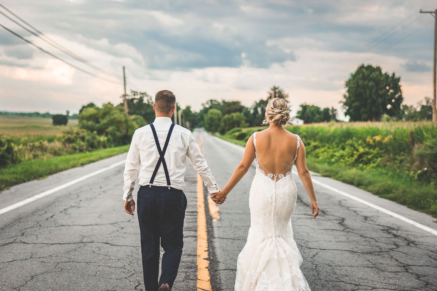 A couple in wedding attire walking down the middle of a country road.