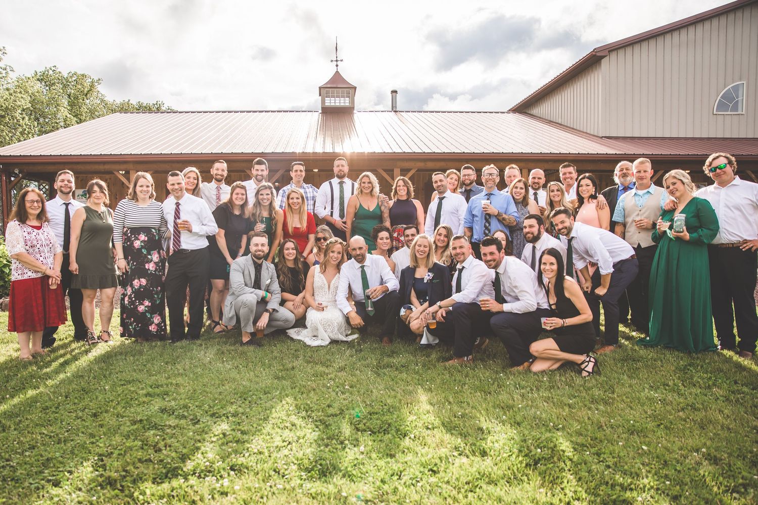 Wedding guests gather on a grassy hillside for an outdoor group photo at a rustic barn venue.