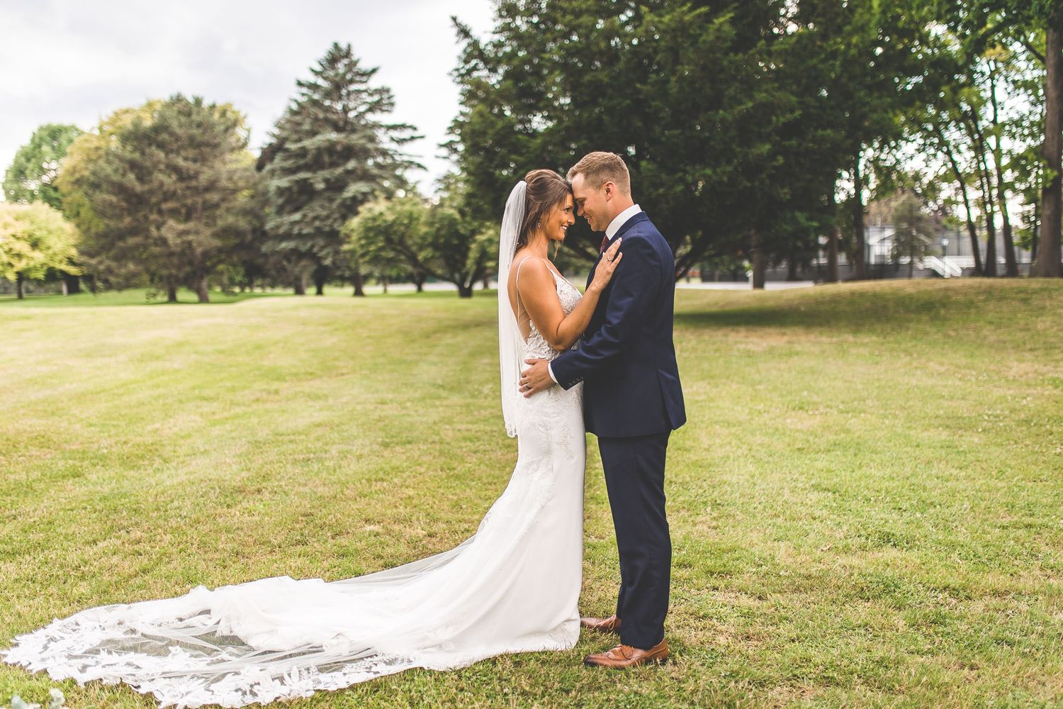 Wedding couple poses together on rolling hills with trees in background.