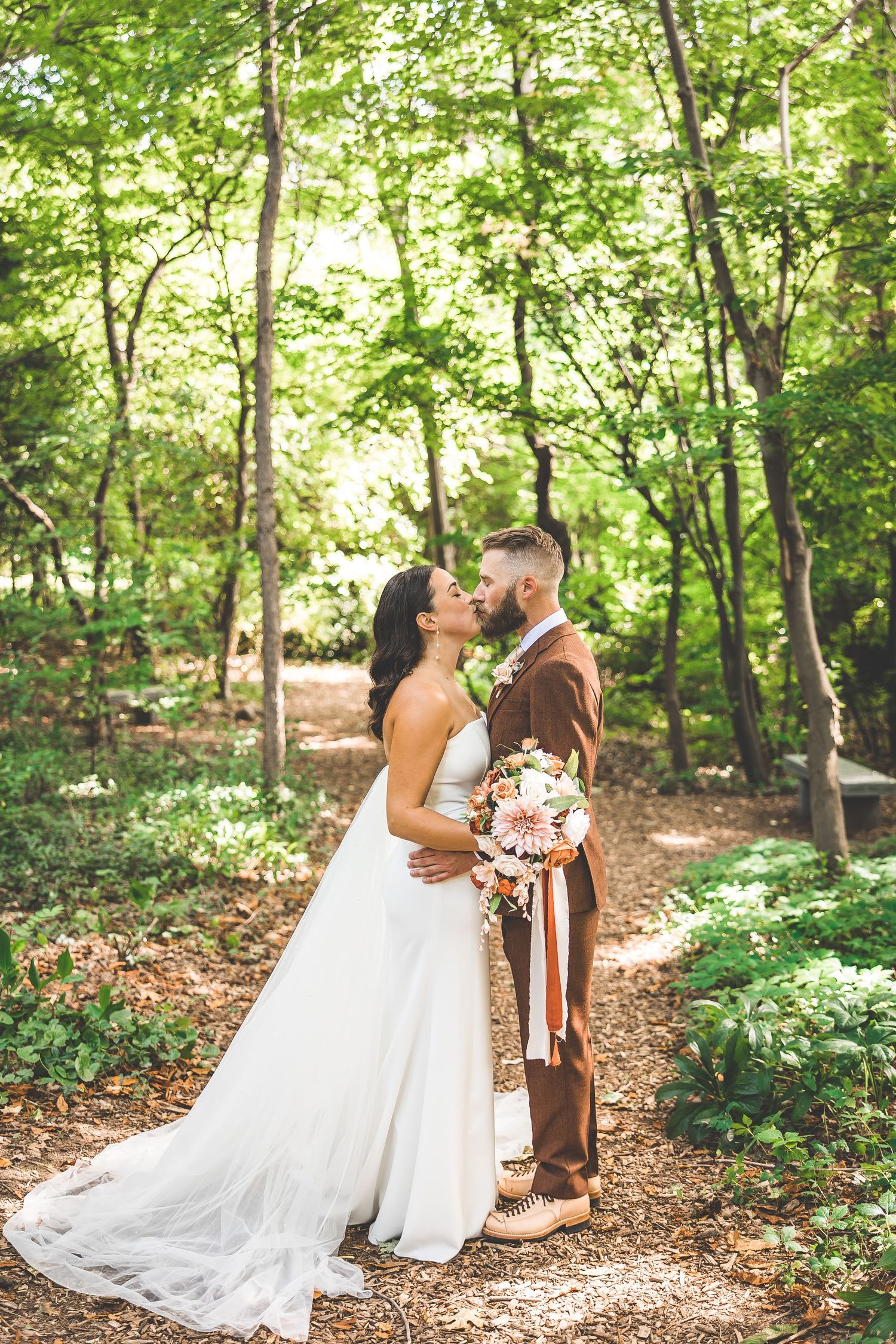 A romantic wedding couple shares an intimate moment on a sunlit forest path surrounded by lush greenery.