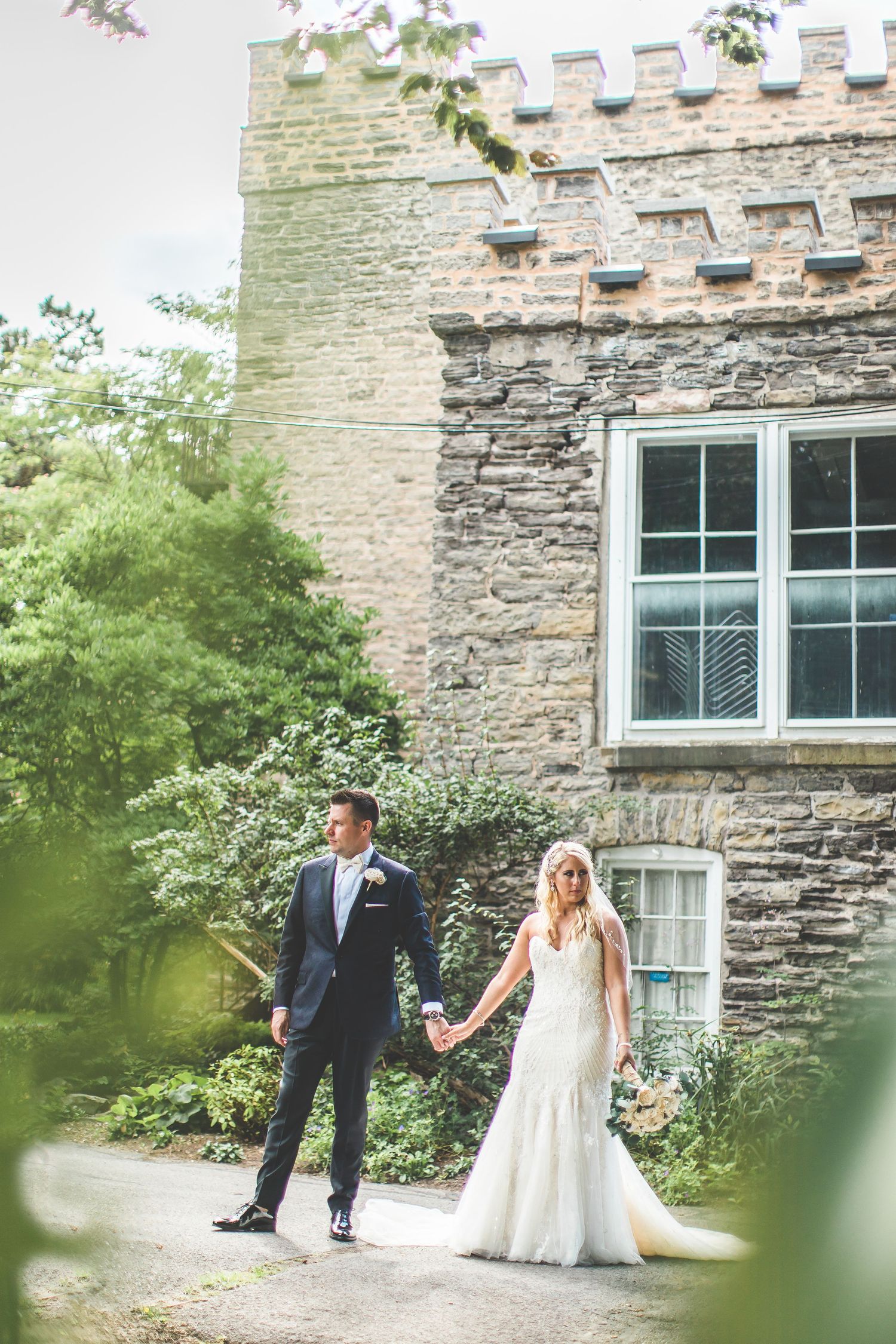 A couple in wedding attire walks hand in hand near a stone castle wall.