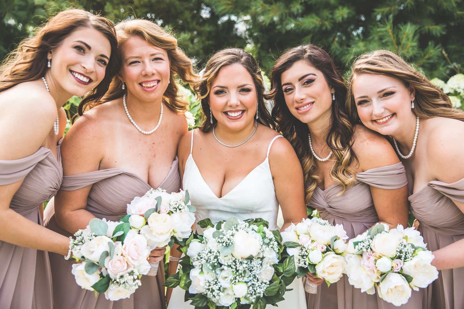 Bridesmaids in neutral dresses holding white floral bouquets pose together outdoors.