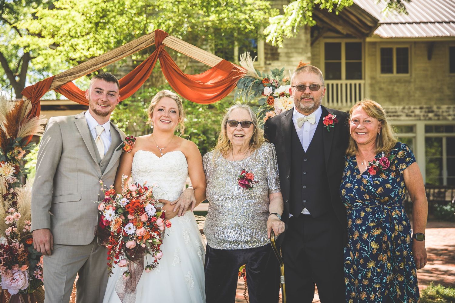 Small wedding group poses together under orange draped arch in garden setting.