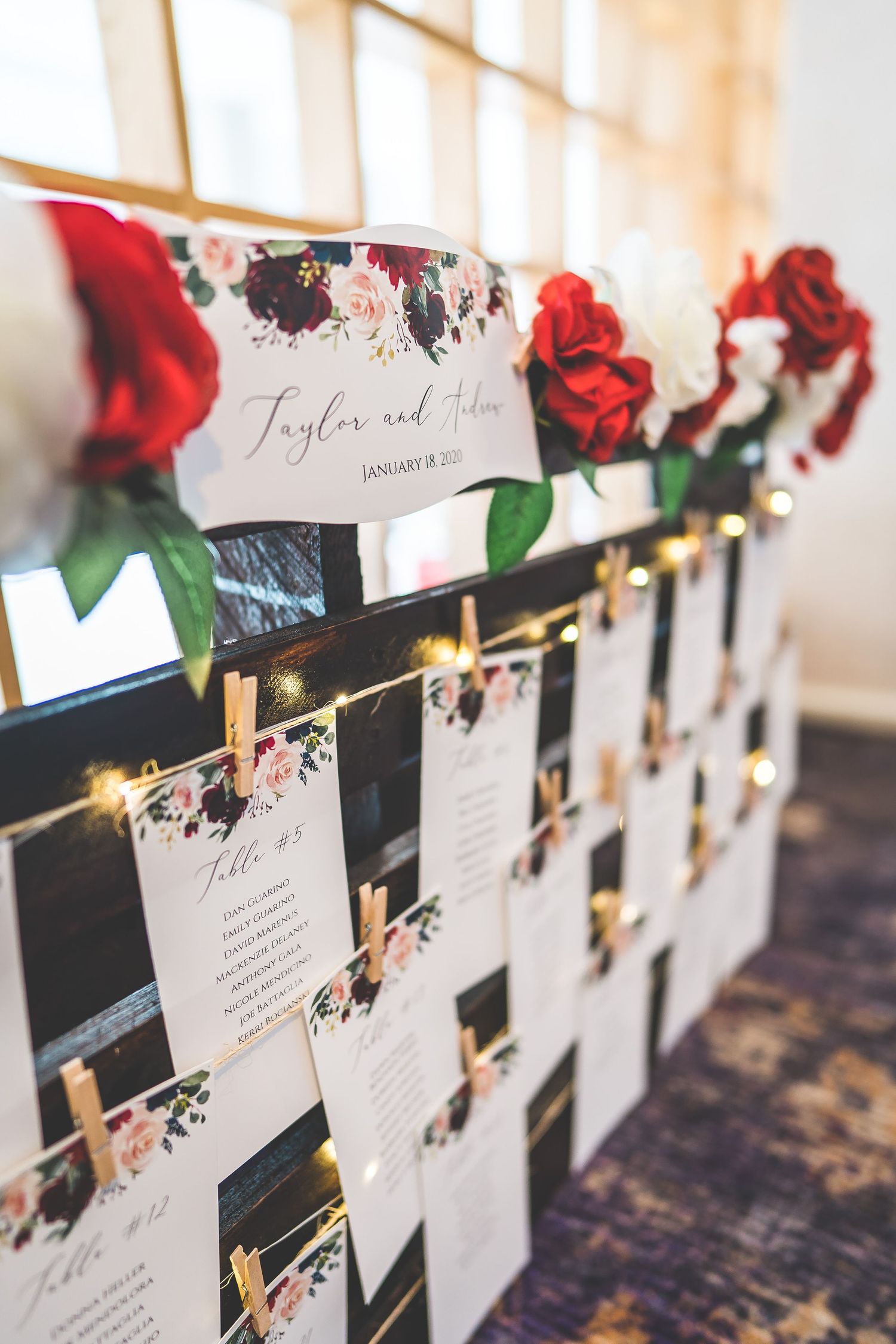 Wedding seating cards decorated with red roses displayed on a string with wooden clothespins.