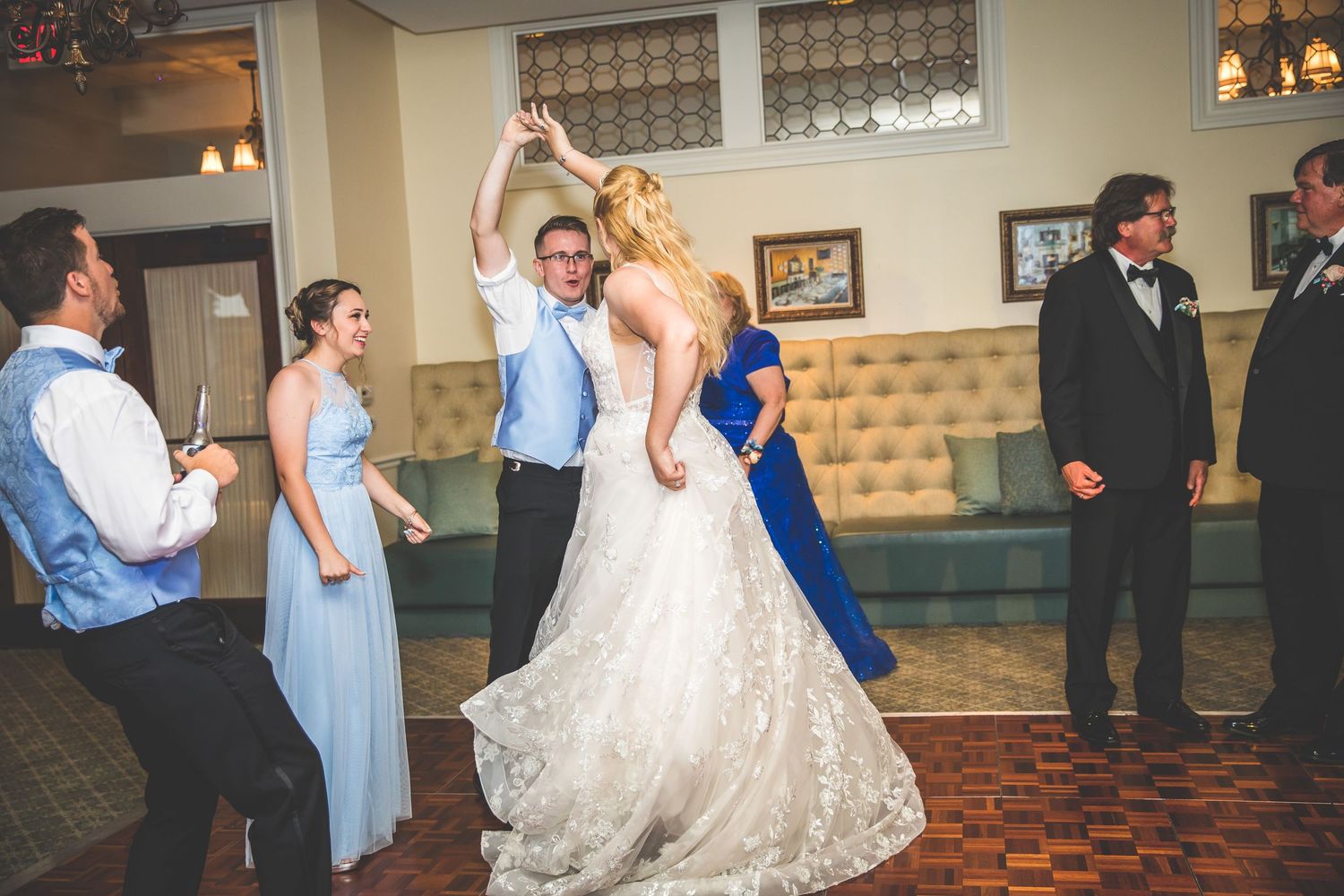 Wedding party members in formal attire dancing on patterned ballroom floor with elegant lighting.