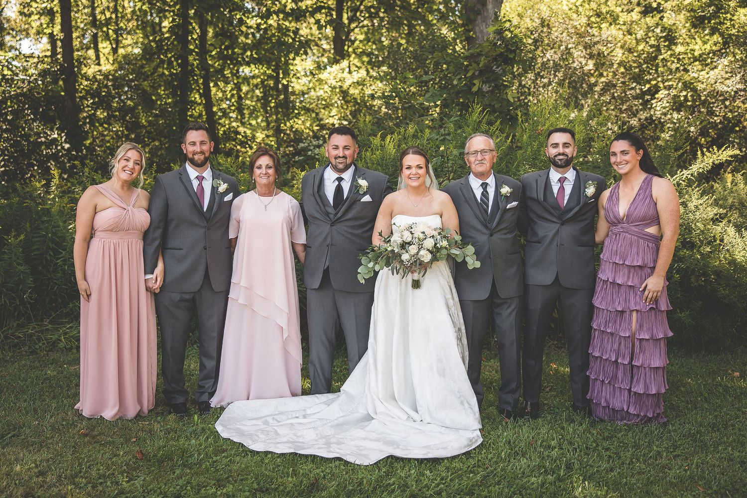 Formal wedding party portrait with bridesmaids in pink and purple dresses against a wooded background.