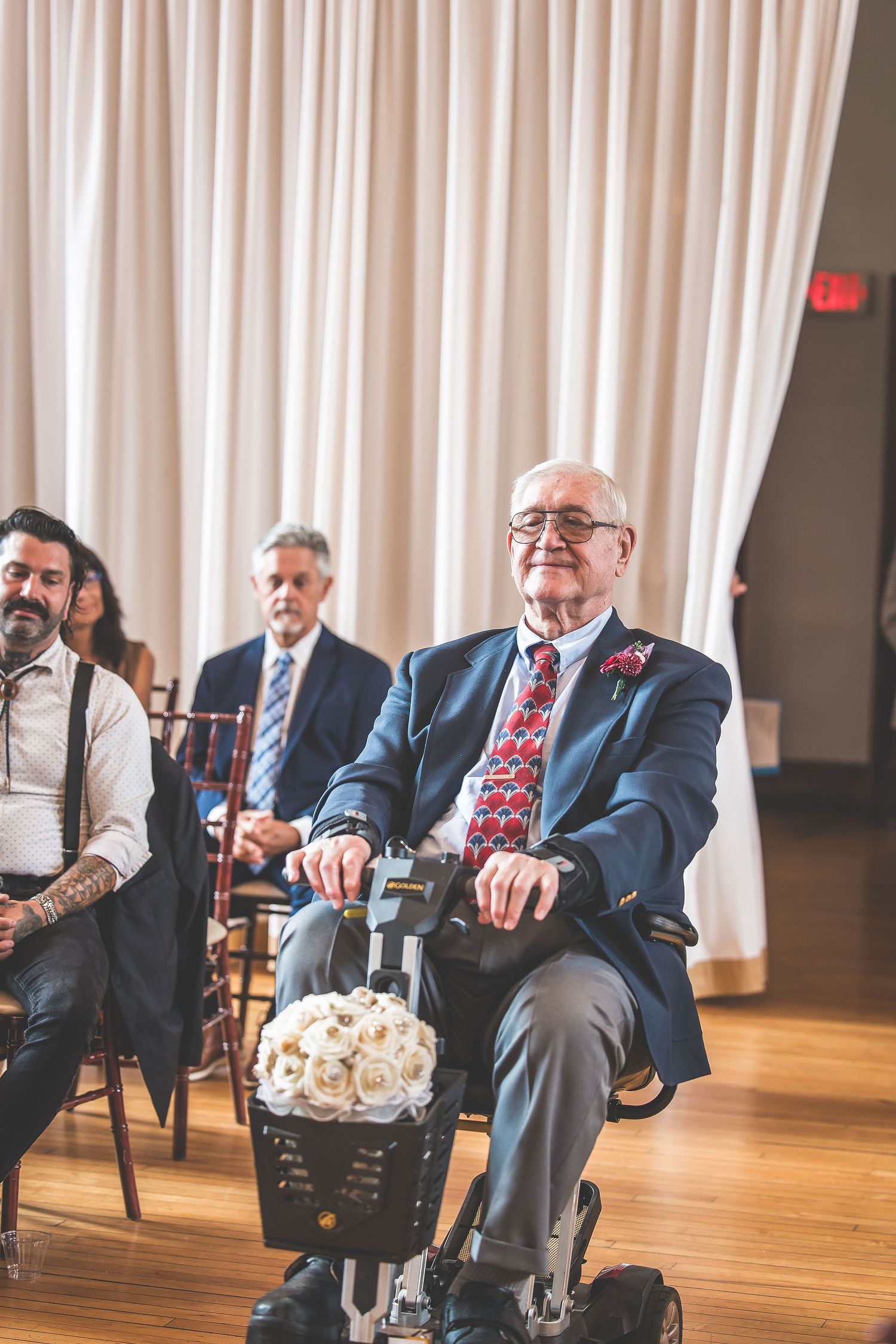 Wedding guests seated in formal attire at an indoor venue with white curtains and hardwood floors.