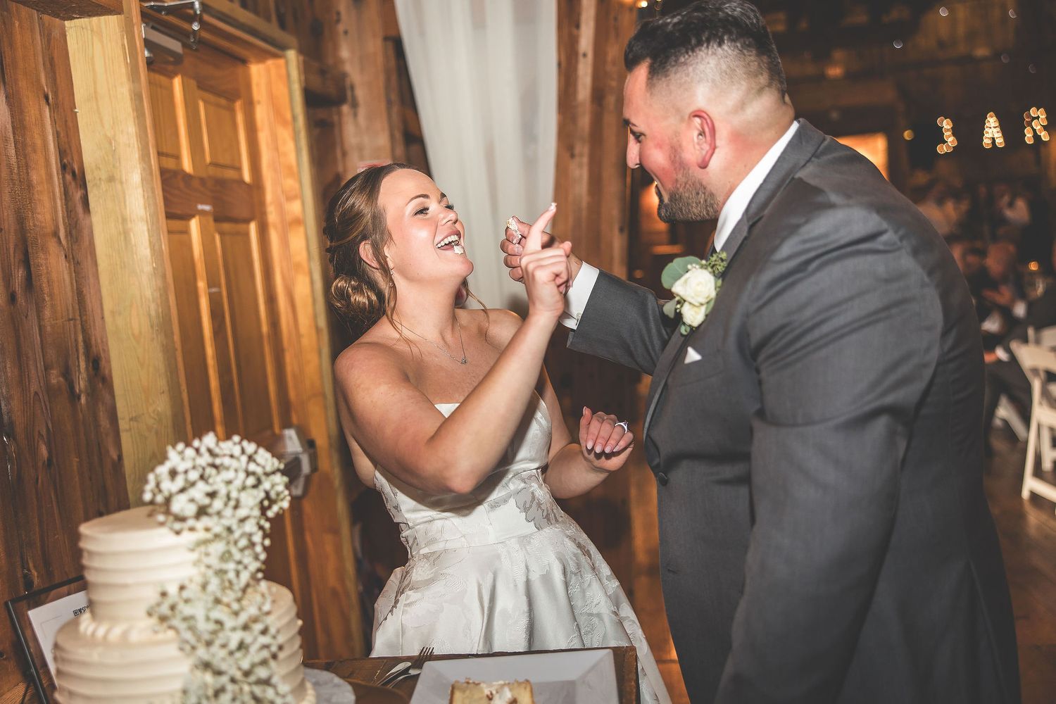 Newlyweds playfully share wedding cake during reception in rustic venue with baby's breath floral decorations.