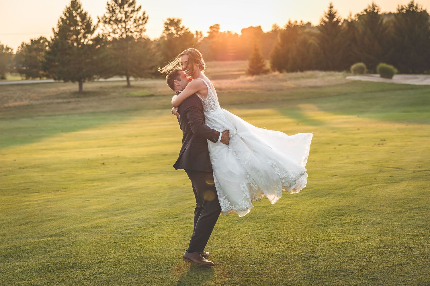 Groom lifts bride in flowing white dress on golf course at sunset.