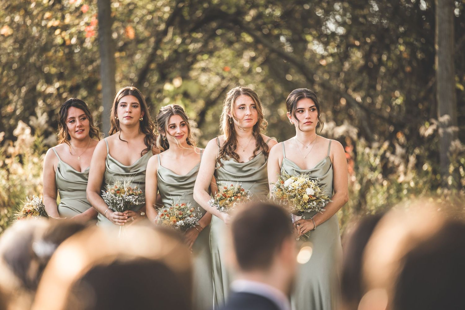 Bridesmaids in sage green dresses holding bouquets during outdoor forest ceremony.