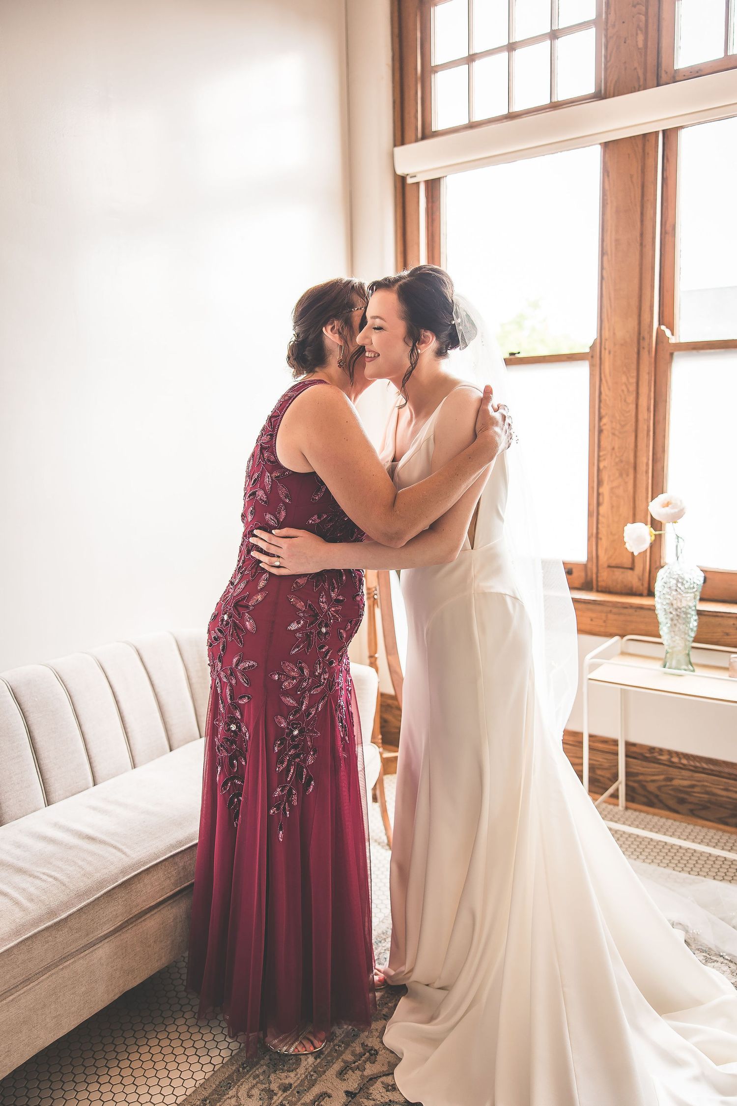 Two people share an intimate moment in front of sunlit windows wearing formal wedding attire.