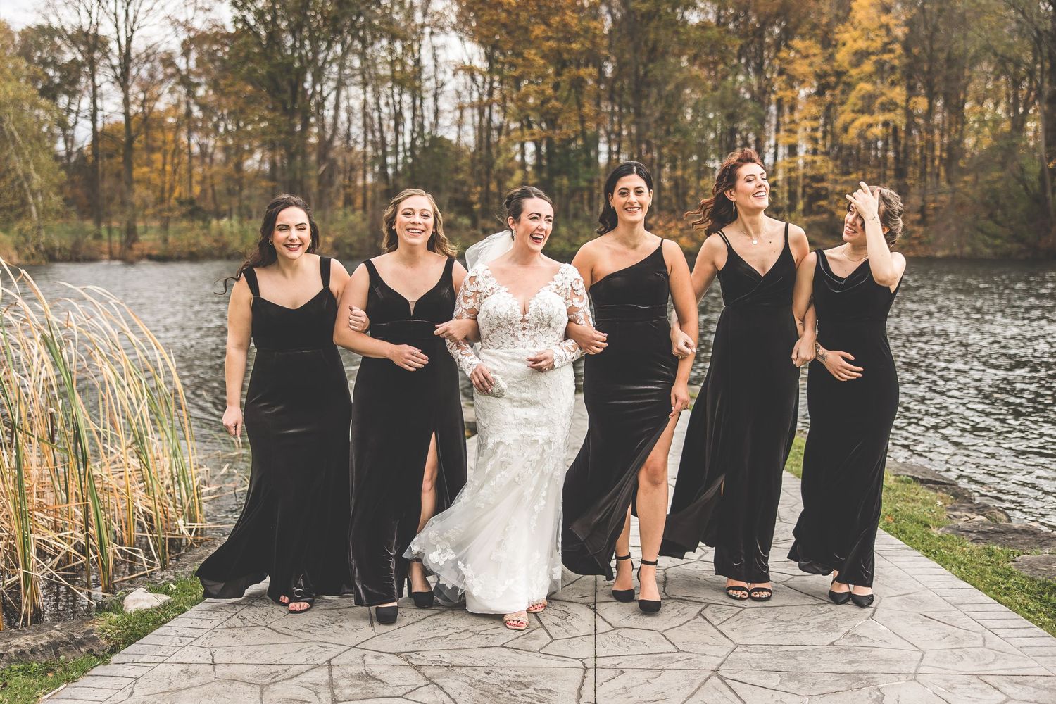 Wedding party laughs together while standing on stone path near autumn lake.